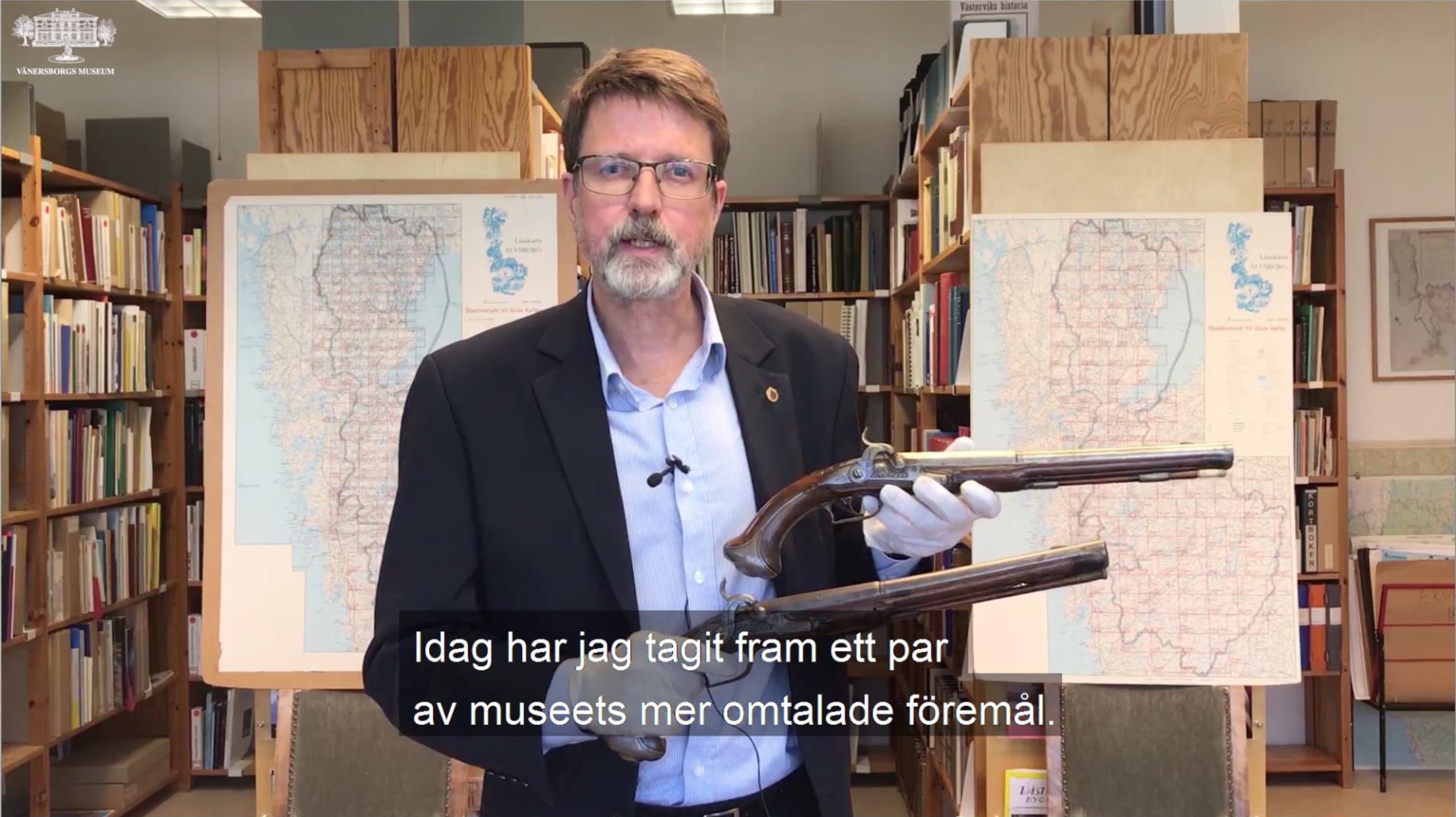 En vit, medelåldersman står i ett magasin flankerad av två kartor, med två pistoler i sina händer.