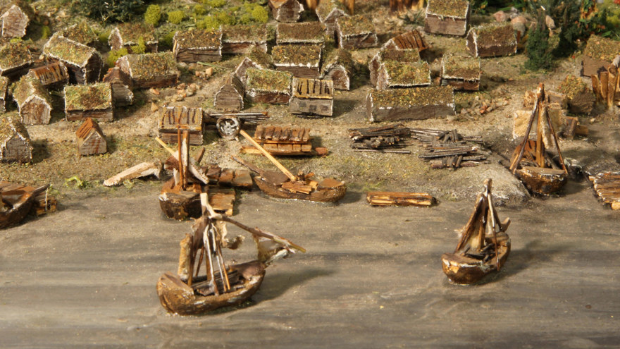 En modell med små hus med mossa på taket vid vattnet där två båtar åker
