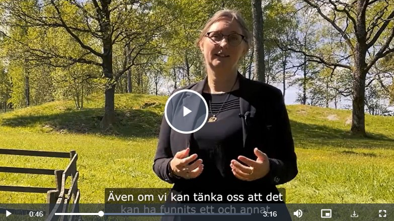 En kvinna med upsatt hår och glasögon berättar. Runt omkring henne syns träd och gräs en vårdag.