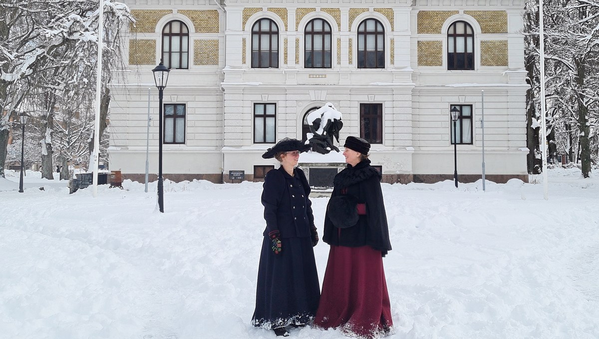 Två kvinnor i 1800-talskläder står i snö framför Vänersborgs museum. 