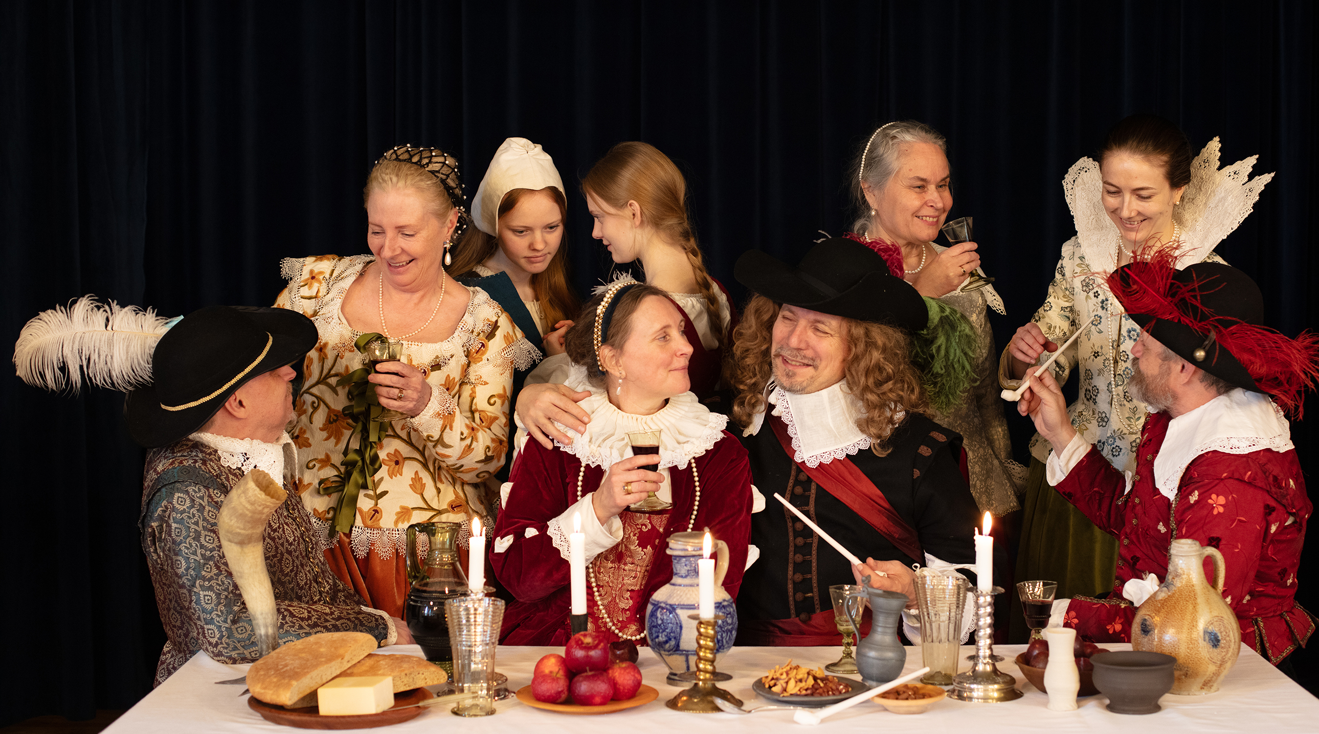 Ett sällskap med människor i tidstyoiska kläder från 1500-talet skålar vid ett dukat bord, de ser ut att tillhöra samhällets mer förmögna